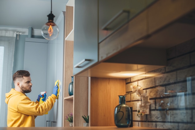 Foto de vista lateral de um homem caucasiano com barba limpando sua casa desinfetando-a com spray usando luvas