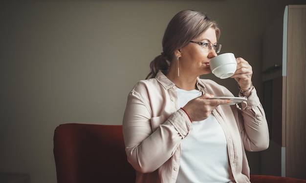 Foto de vista lateral de uma mulher idosa bebendo uma xícara de chá enquanto olha pela janela de dentro de casa
