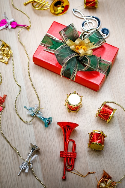 Foto de vista superior da caixa de presente embrulhada em papel vermelho pequeno com gravata borboleta de fita brilhante verde e dourada colocada na mesa de madeira com pequenos instrumentos musicais decorativos na véspera de Natal ou festival de ano novo.