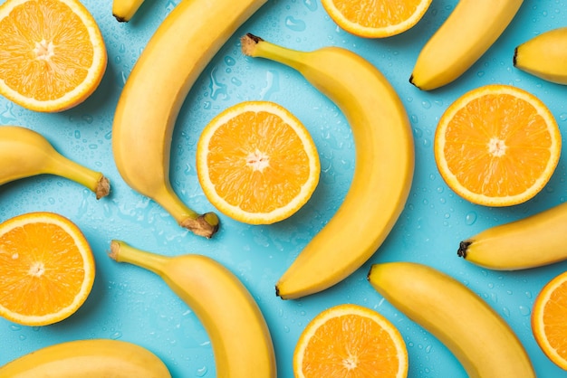 Foto de vista superior de gotas de água espalhadas bananas com casca e laranjas cortadas em fundo azul pastel isolado