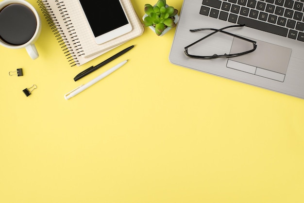 Foto de vista superior de óculos de laptop planta canetas fichário prende xícara de bebida e celular em dois lembretes em espiral em fundo amarelo pastel isolado com espaço em branco