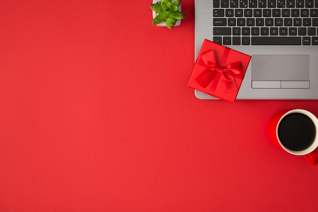 Foto de vista superior de pequena caixa de presente vermelha vívida com laço de fita vermelha na planta do laptop e xícara de bebida em fundo vermelho isolado com copyspace