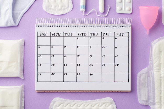 Foto de vista superior do calendário lingerie rosa copo menstrual pílulas analgésicas almofadas higiênicas e tampões em fundo lilás pastel isolado