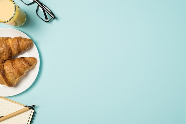 Foto de vista superior do copo de óculos no local de trabalho de caderno de caneta de suco e prato com dois croissants em fundo azul pastel isolado com espaço vazio