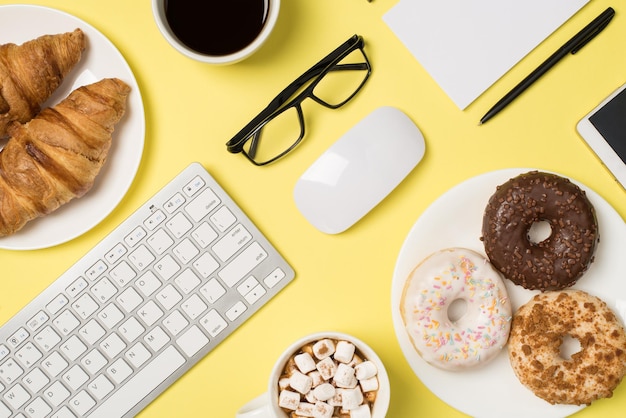 Foto de vista superior do teclado mouse óculos caneta caderno duas xícaras de bebida com marshmallow e café dois pratos com croissants e rosquinhas e smartphone em fundo amarelo pastel isolado