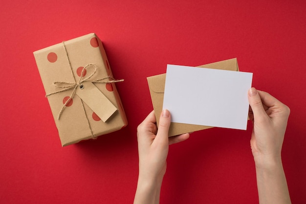 Foto de vista superior em primeira pessoa de mãos de decoração de dia dos namorados de santo segurando folha de papel e envelope sobre caixa de presente com padrão de bolinhas e laço de barbante em fundo vermelho isolado com espaço em branco