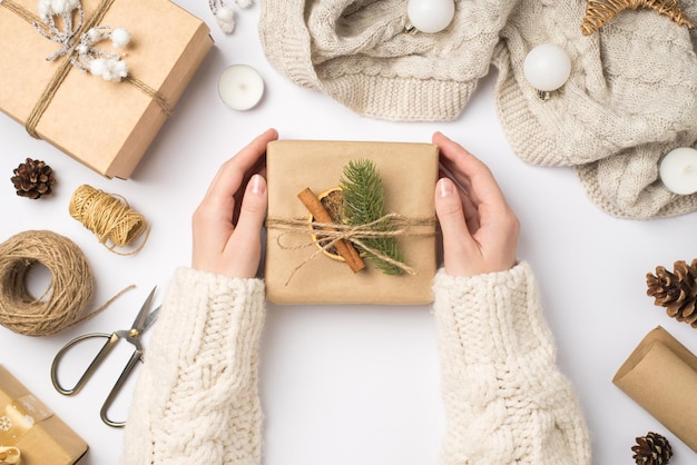 Foto de vista superior em primeira pessoa de mãos no suéter segurando caixa de presente de papel artesanal com laço de pinheiro galho de canela pau de canela fatia de limão seca cachecol bolas e ferramentas para artesanato em fundo branco isolado