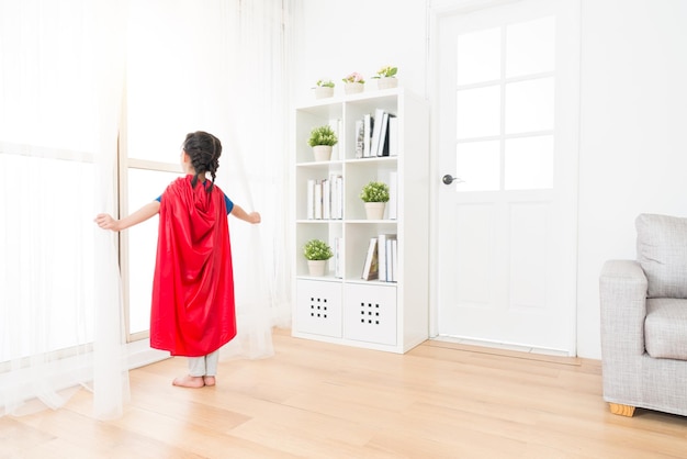 foto de vista traseira da juventude adorável garotinha crianças brincando como super-herói e de pé na sala de estar, abrindo a cortina do chão de madeira, olhando para a janela.