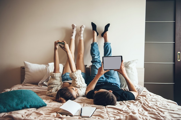 Foto de vista traseira de um casal deitado na cama com tecnologias modernas e sorrindo um para o outro, posando perto de um livro