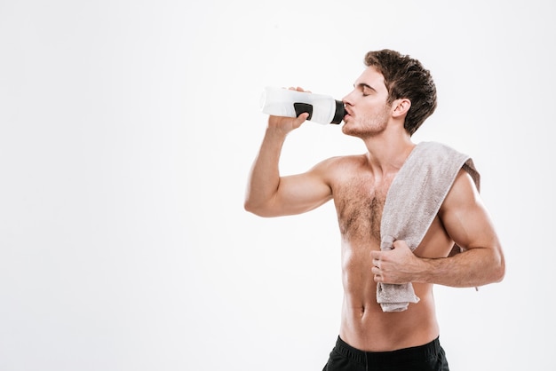 Foto de deportista guapo de pie con una toalla sosteniendo una botella de agua mientras bebe sobre una pared blanca.