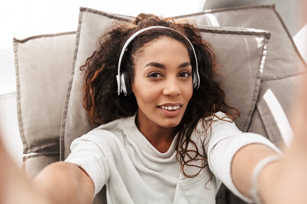 Foto der attraktiven Afroamerikanerfrau, die Kopfhörer trägt, die selfie auf Handy nehmen, während auf Sofa in heller Wohnung liegend