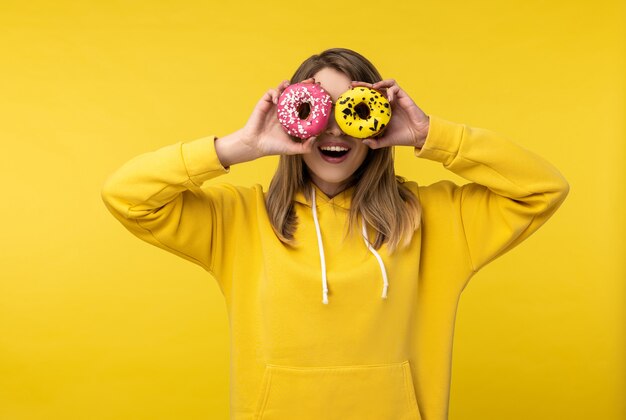 Foto der attraktiven Dame hält Donuts und bedeckte Augen und lächelt. Trägt lässigen gelben Kapuzenpulli, isolierten gelben Farbhintergrund.