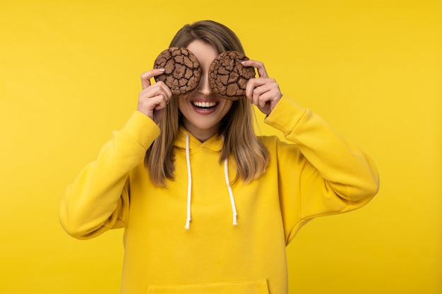 Foto der attraktiven Dame hält Kekse und deckt die Augen ab. Trägt lässigen gelben Kapuzenpulli, isolierten gelben Farbhintergrund.