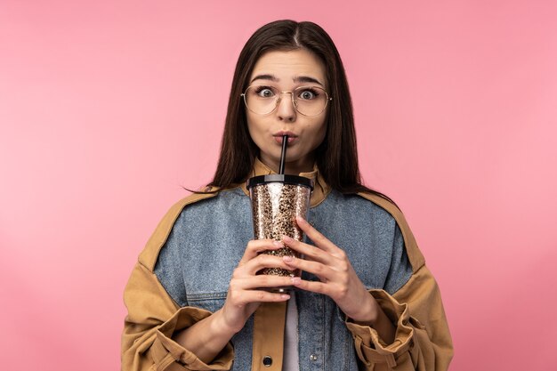 Foto der attraktiven Dame in den Gläsern hält trinken Tee Kaffee und schockiert, trägt lässige Jeansjacke weißes Unterhemd lokalisierten rosa Farbhintergrund.