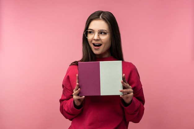 Foto der attraktiven Dame in Gläsern hält Schachtel mit Geschenk, wartet und freut sich zu öffnen. Trägt lässige rosa Pullover weiße Hosen isoliert rosa Farbe Hintergrund.
