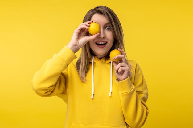 Foto der attraktiven Dame posiert auf niedliche Weise mit Zitronen, bedecken Sie ein Auge mit. Trägt lässigen gelben Kapuzenpulli, isolierten gelben Farbhintergrund.