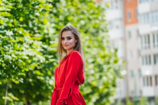 Foto der attraktiven Frau im roten Kleid, das nahe grünem hellem Baum steht. Frühling Herbst. Junges Mädchen in hellem Licht und grünen Blättern. Unscharfer Hintergrund. Nahaufnahme.