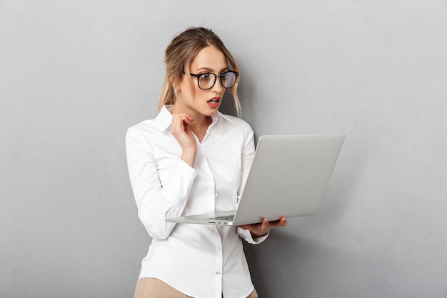 Foto der attraktiven Geschäftsfrau, die die Brille steht und Laptop im Büro hält, lokalisiert