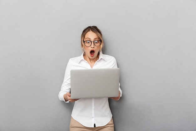 Foto der aufgeregten sachlichen Frau, die Brille steht und Laptop im Büro hält, lokalisiert