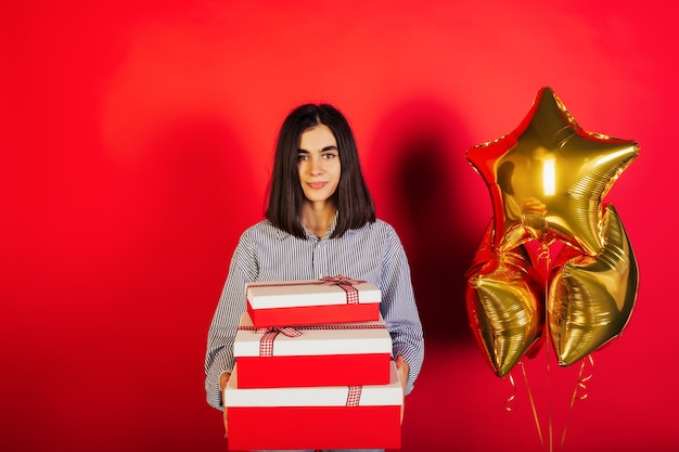 Foto der Dame, die drei große Geschenkboxen und drei goldene Luftballons hält, lokalisiert über rotem Farbhintergrund. Geburtstagsparty.