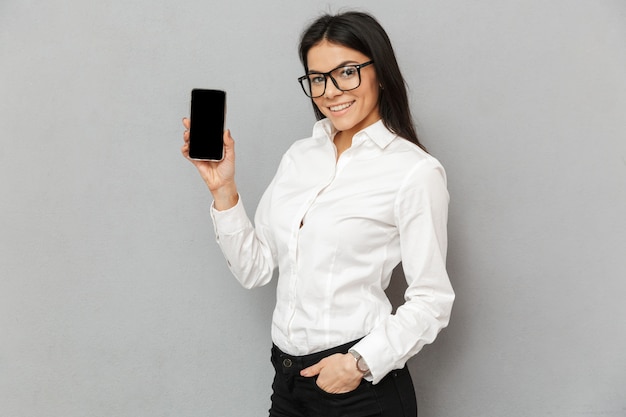 Foto der eleganten jungen Frau 30 in formeller Kleidung, die mit Smartphone in den Händen steht und schwarzen Bildschirm auf Kamera demonstriert, lokalisiert über grauem Hintergrund