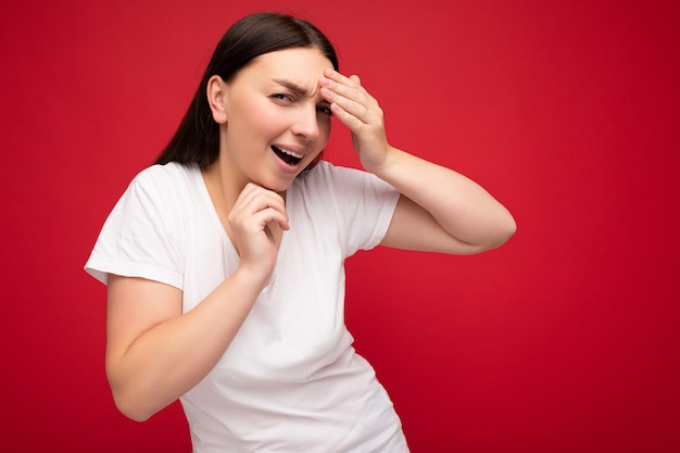 Foto der emotionalen jungen schönen brünetten Frau mit aufrichtigen Gefühlen, die weißes T-Shirt für Modell lokalisiert über rotem Hintergrund mit leerem Raum und berührendem Kopf tragen.