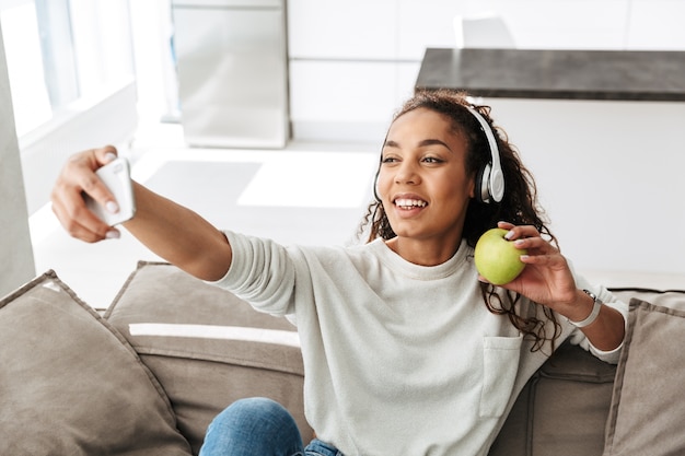 Foto der erfreuten Afroamerikanerfrau, die Kopfhörer trägt, die selfie auf Handy nehmen, während auf Couch in heller Wohnung sitzend