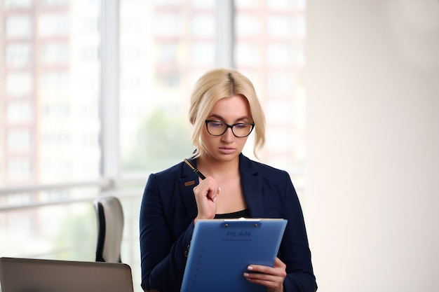 Foto der ernsten Manager- oder Direktorfrau, die formelle Kleidung und Brillen trägt, die in den Papierdokumenten der Hände halten, während sie im Büro auf Laptop arbeiten