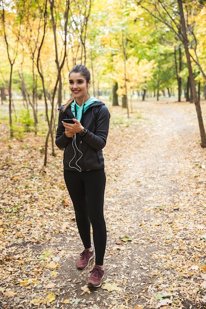 Foto der erstaunlichen jungen hübschen Fitnessfrau draußen im Park unter Verwendung des Mobiltelefons, das Musik mit Kopfhörern hört.