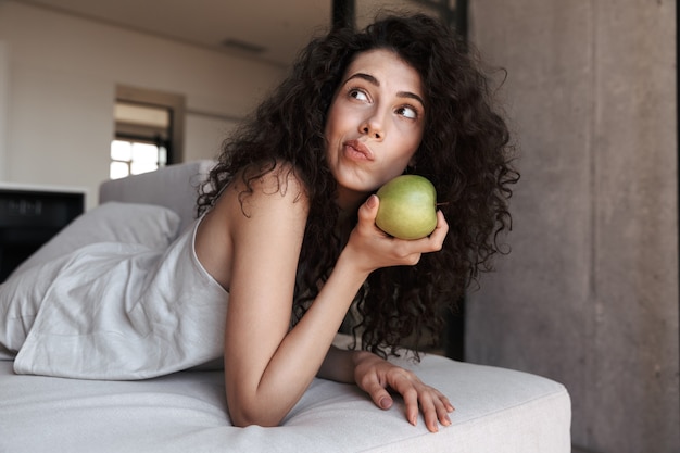 Foto der europäischen lockigen Frau mit dem langen dunklen Haar, das Freizeitkleidung der Seide trägt, die mit Interesse beiseite schaut, während großen grünen Apfel während der Ruhe auf Sofa in der Wohnung hält
