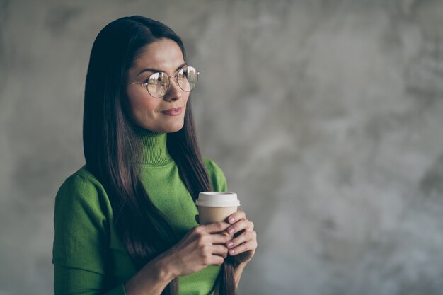 Foto der fröhlichen Genießfrau, die heißen Kaffee vom Papierbecher trinkt, der weg in den Brillen lokalisierten grauen Farbwandbetonhintergrund schaut