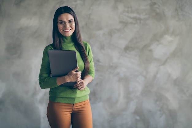 Foto der fröhlichen positiven hübschen Frau, die Laptop mit den Händen zahnig lokalisiert nahe leerem Raum durch grauen Wandbetonhintergrund hält