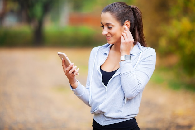 Foto der frohen Eignungs-Frau 30s in der Sportkleidung, die Bluetooth Earpod berührt und Handy hält, beim Stillstehen im grünen Park