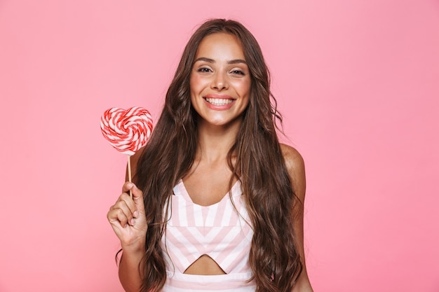 Foto der glücklichen Frau 20s, die Kleid lacht und große Süßigkeiten isst, lokalisiert über rosa Wand