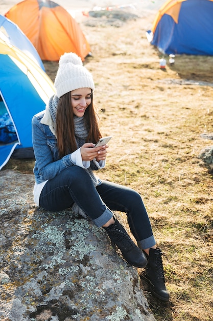 Foto der glücklichen jungen Frau draußen mit Zelt im freien alternativen Feriencamping über den Bergen mit Handy.