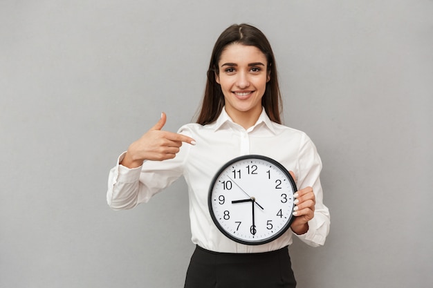 Foto der glücklichen jungen Frau im weißen Hemd und im schwarzen Rock, der Finger auf der großen runden Uhr zeigt, die in der Hand hält, lokalisiert über graue Wand