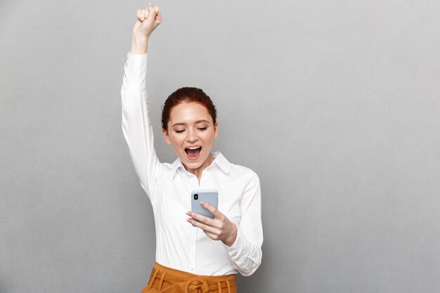 Foto der glücklichen jungen hübschen rothaarigen Geschäftsfrau, die lokalisiert über grauer Wand unter Verwendung des Handys aufwirft, das Siegergeste zeigt.