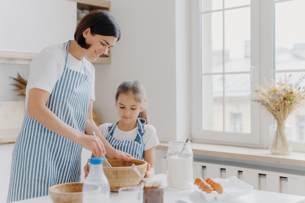 Foto der glücklichen Mutter und des Kindes, die zusammen in der Küche kochen, tragen Schürzen, etwas Leckeres zubereiten, Essen zubereiten, Eier mit Schneebesen verquirlen, Hausgebäck zu Hause backen, Milcheier, Schokoladenmehl auf dem Tisch