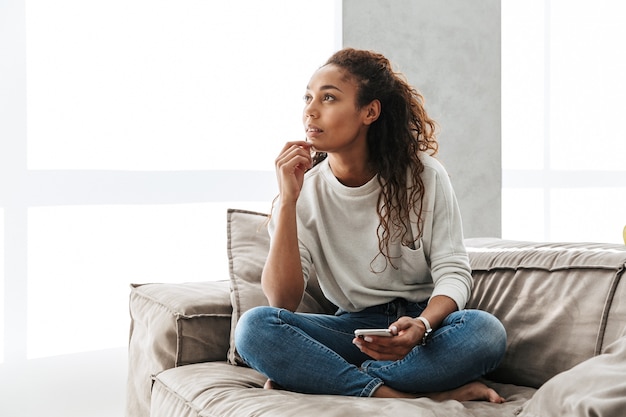 Foto der hübschen Afroamerikanerfrau, die Handy benutzt, während sie auf Couch in heller Wohnung sitzt