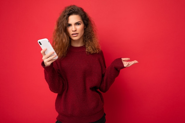 Foto der hübschen fragenden verärgerten jungen Frau mit dem lockigen Haar, das dunkelroten Pullover trägt