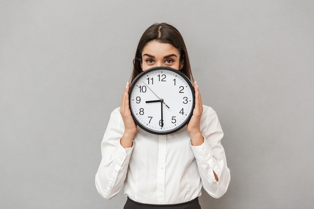 Foto der hübschen Geschäftsfrau in der formellen Kleidung, die Gesicht mit großer runder Uhr bedeckt, lokalisiert über graue Wand