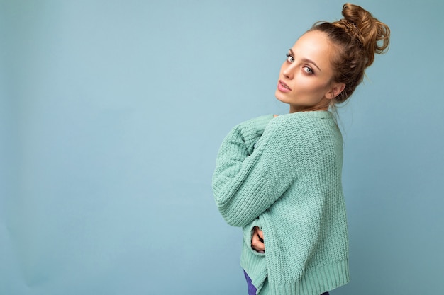 Foto der jungen attraktiven europäischen Modeblondinenfrau, die blauen Pullover einzeln über blauem Hintergrund mit Kopienraum mit ernstem Gesichtsausdruck trägt. Einfache und natürliche weibliche Person