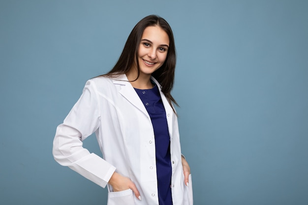 Foto der jungen attraktiven schönen positiven positiv lächelnden Brünettenfrau mit aufrichtigen Gefühlen, die weißen medizinischen Mantel tragen, lokalisiert über blaue Wand mit leerem Raum.