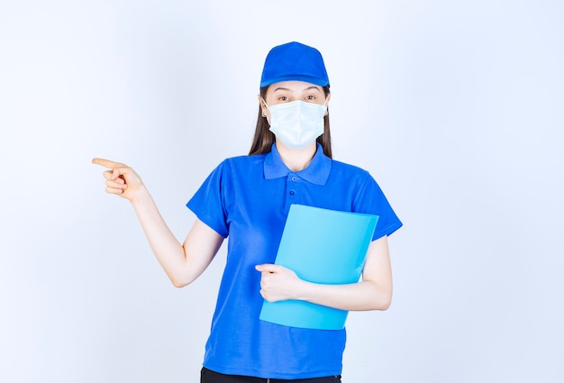 Foto der jungen Frau in medizinischer Maske, die Ordner hält und mit dem Finger beiseite zeigt.