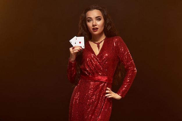 Foto der jungen schönen kaukasischen brünetten Frau im glänzenden roten Abendkleid hält Karten in einer Hand, zeigt ein Paar Asse und hält eine Hand auf der Hüfte. Brauner Hintergrund, Casino, Glücksspielindustrie