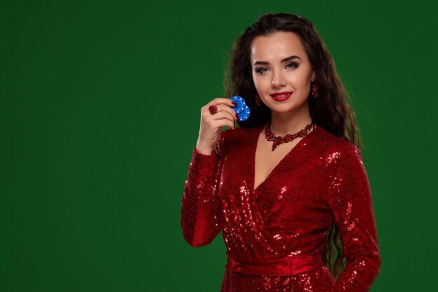 Foto der jungen schönen sexy glücklichen Brunettefrau in einem glänzenden Kleid des roten Abends hält einige Schürhakenchips in ihren Händen. Grüner Hintergrund, Chromakey, Glücksspielindustrie