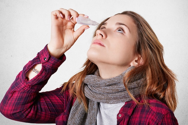 Foto der kranken Frau hat schlechtes Sehvermögen, verwendet Spray für tropfende Augen, trägt gestrickten Schal, isoliert über weißer Wand. Kranke Frau benutzt Aerosal Spray. Konzept für Medizin, Gesundheitswesen und Krankheit