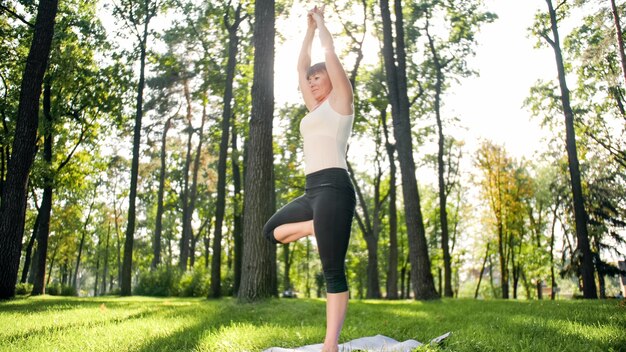 Foto der lächelnden glücklichen Frau 40 Jahre alt, die Yogaübungen auf Fitnessmatte im Wald macht. Harmonie des Menschen in der Natur. Menschen mittleren Alters, die sich um die geistige und körperliche Gesundheit kümmern