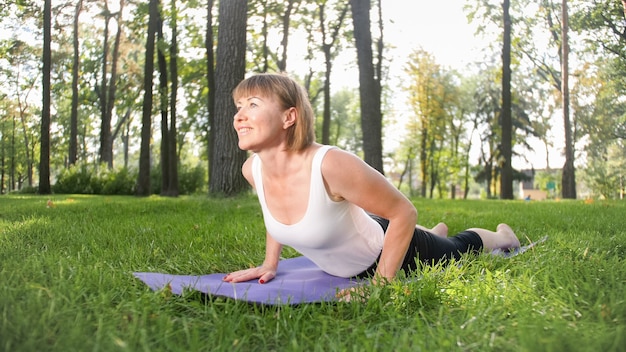 Foto der lächelnden glücklichen Frau 40 Jahre alt, die Yogaübungen auf Fitnessmatte im Wald macht. Harmonie des Menschen in der Natur. Menschen mittleren Alters, die sich um die geistige und körperliche Gesundheit kümmern