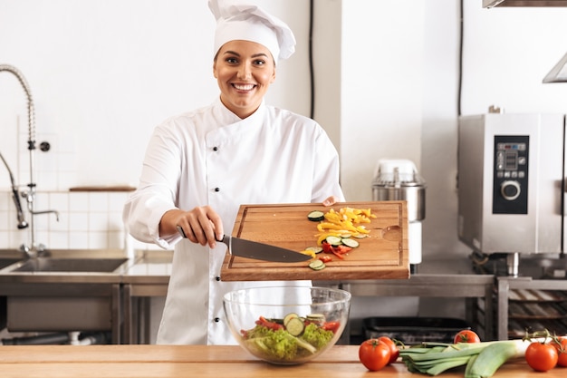 Foto der lächelnden Köchin, die weiße Uniform trägt, die Salat mit frischem Gemüse macht, in der Küche am Restaurant